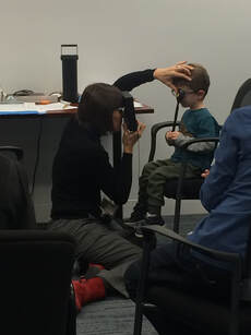 An optometrist kneels on the floor to examine a child's left eye. The child holds an occluder over their right eye. 